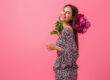 Una mujer sonriente sostiene un ramo de flores, reflejando felicidad, feminidad y frescura. Con un fondo rosa vibrante, la imagen transmite positividad, belleza y empoderamiento femenino.