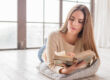 Mujer joven leyendo un libro antiguo mientras descansa sobre un cojín, en una habitación iluminada por luz natural.