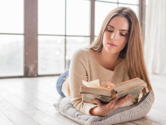 Mujer joven leyendo un libro antiguo mientras descansa sobre un cojín, en una habitación iluminada por luz natural.